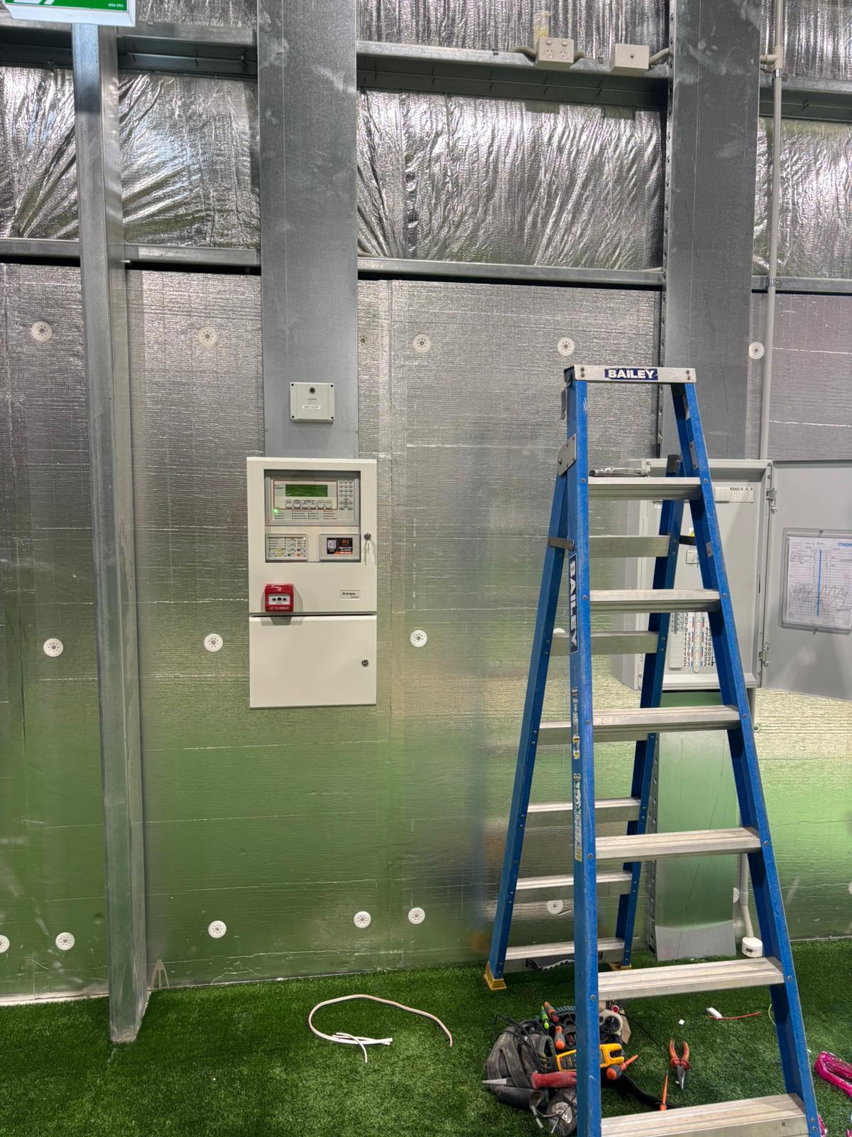 Freshly installed fire detection panel inside a sports facility with ladder and tools